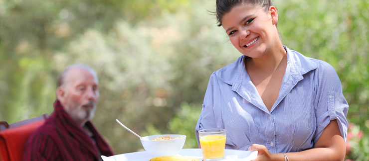 Female caregiver serves breakfast outside to elderly man sitting at table outside.