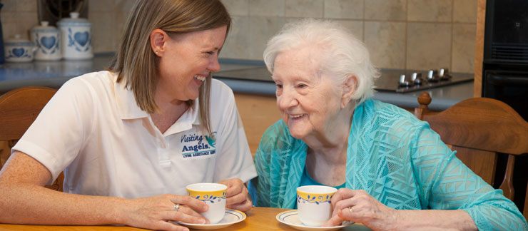 Female care aide smiles while drinking cup of tea with elderly woman at the kitchen table.