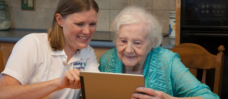 Female caregiver helps elderly woman looking at a computer tablet while sitting at the kitchen table.