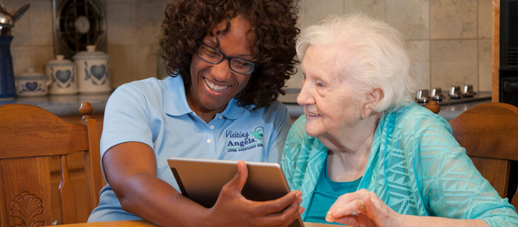 Female caregiver smiles while showing computer tablet to elderly woman at kitchen table.