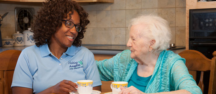 A female in-home care provider in a blue collared shirt sits at a table next to an elderly woman in a turqiose blouse. Both are smiling at each other and enjoying a cup of tea. 