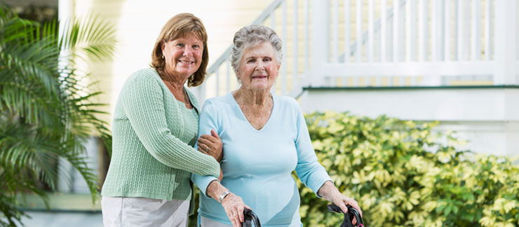 Female caregiver holds the arm of an elderly woman with walker outside.
