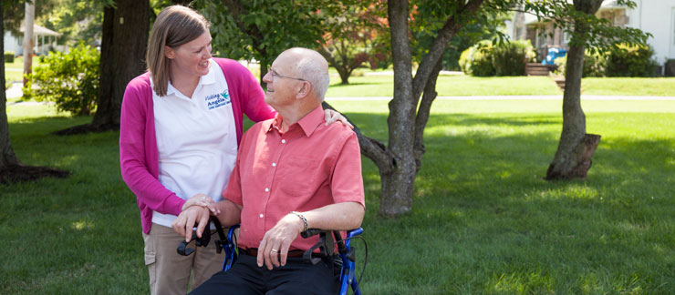 An in-home care nurse in a Visiting Angels polo and pink sweater stands next to an elderly man seated in a blue walker. Her arm is around his shoulders