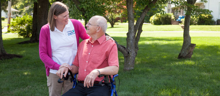 Female care worker smiles next to elderly man sitting in a wheelchair outside his home.