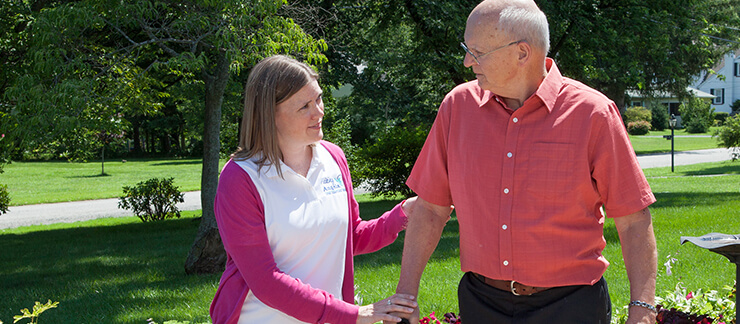 Female care worker places hand on the arm of senior man standing outside.