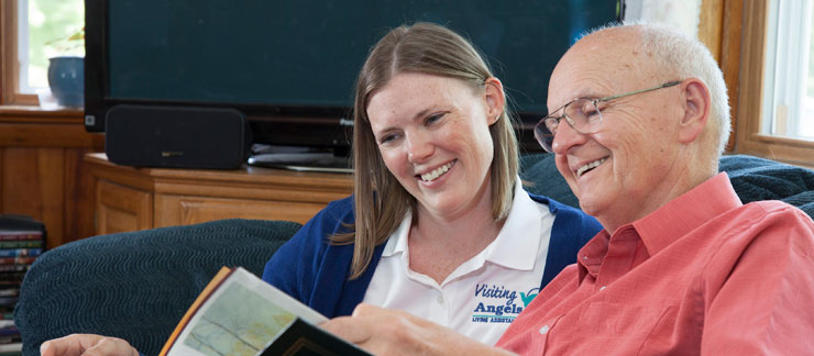 Female caregiver smiles while looking at a book with elderly man on couch.