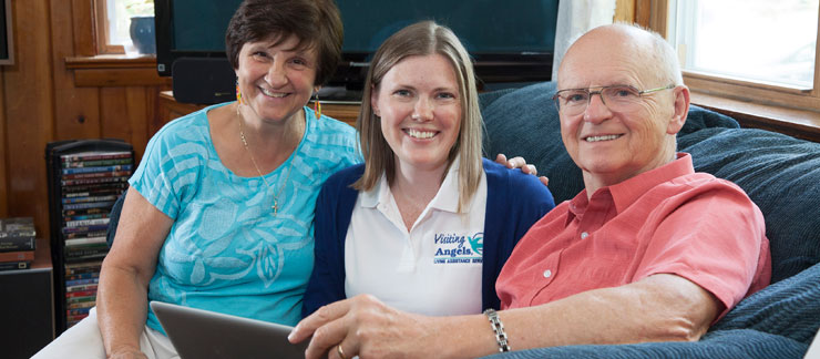 Home care worker sits on couch with smiling elderly couple inside their home.