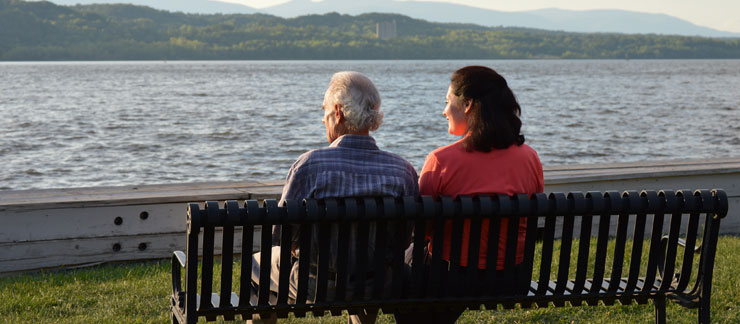 Female caregiver sits with an elderly man on a bench overlooking a lake.
