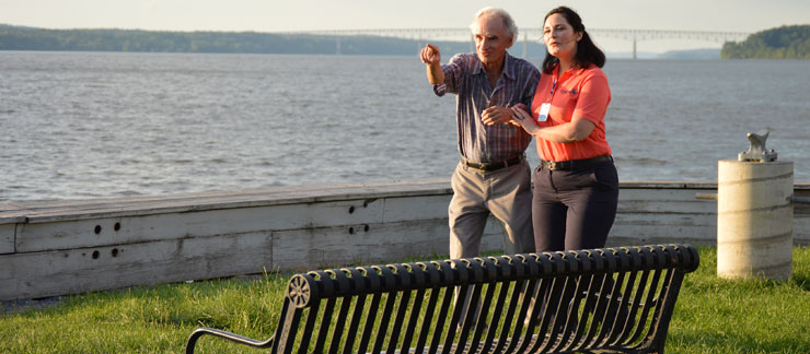 Female home care worker holds the arm of an elderly man to make sure he doesn't get lost as they walk along a path near the river.