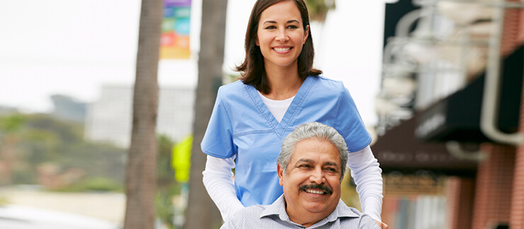 A female in-home care provider in blue scubs pushing an elderly man in his wheelchair down a business and tree-lined sidewalk