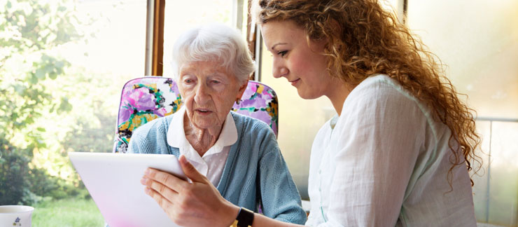 Female caregiver shows an online app on a computer tablet to a senior woman sitting on a chair at home.