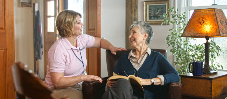 Female home care provider kneels down to talk to elderly woman reading book on couch at home.