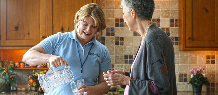 Female home caregiver pours water from pitcher into a glass for elderly woman in kitchen.