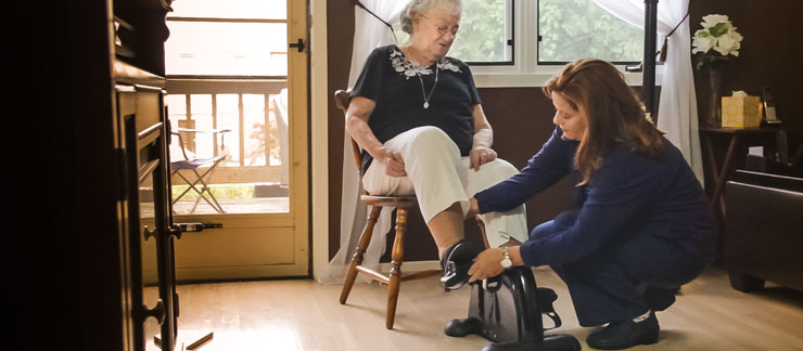 An in-home care provider in navy blue scrubs kneels down to assist a sitting elderly woman peddaling on a stationary leg machine