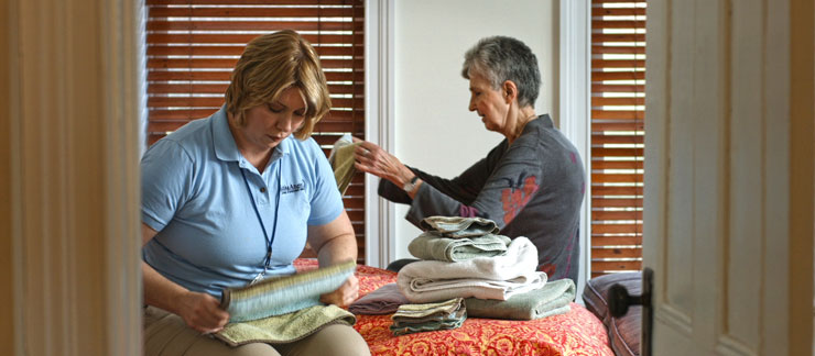 Female care worker helps elderly woman fold clothes in bedroom of home.