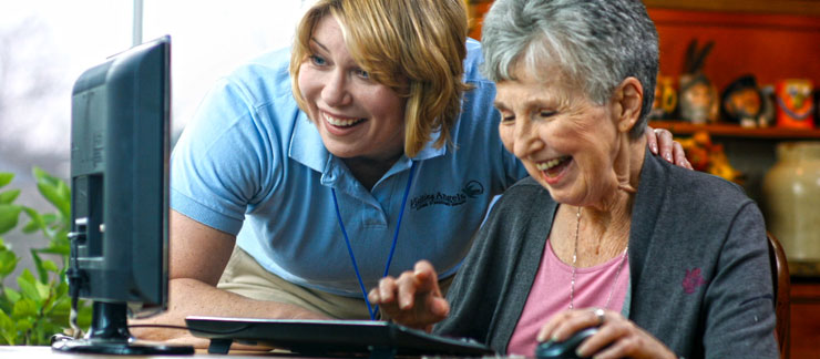 Female care aide assists smiling senior woman working on a computer at home.