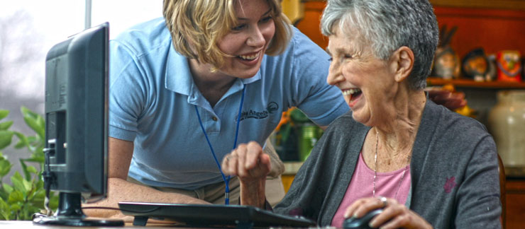 A female in-home care nurse leans over smiling with her hand on an elderly woman's shoulder who is sitting at a desk and smiling at a computer monitor