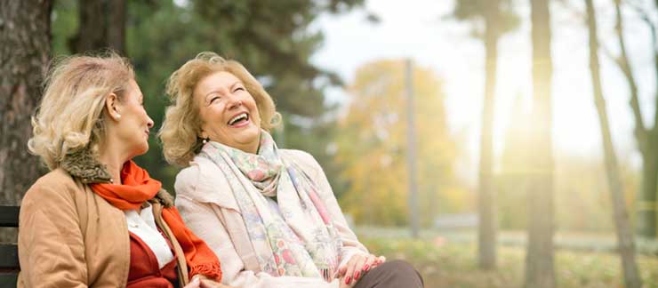 Adult daughter and senior mother laugh while sitting on a park bench in the afternoon.