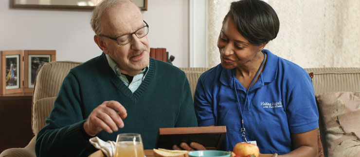 Female caregiver looks at an old photo with elderly man sitting on couch. 