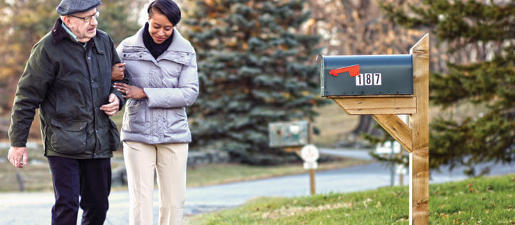 Elderly man in winter coat gets assisted walking from a female caregiver outside of his home near a mailbox.