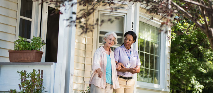 An in-home care provider and elderly woman are walking in front of a home's porch interlocked at the elbow 
