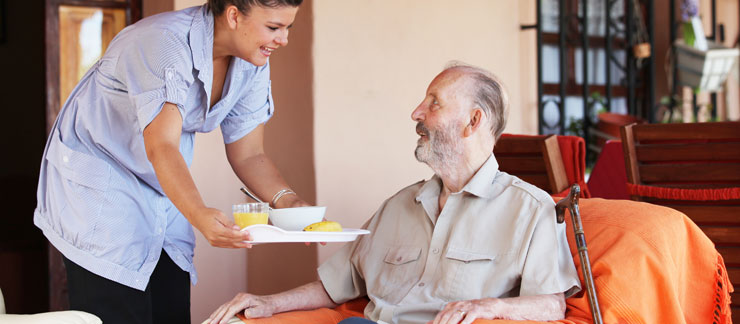 An in-home care provider stands crouched next to an elderly man who is seated as she extends a tray of food to him. The two are smiling at each other.