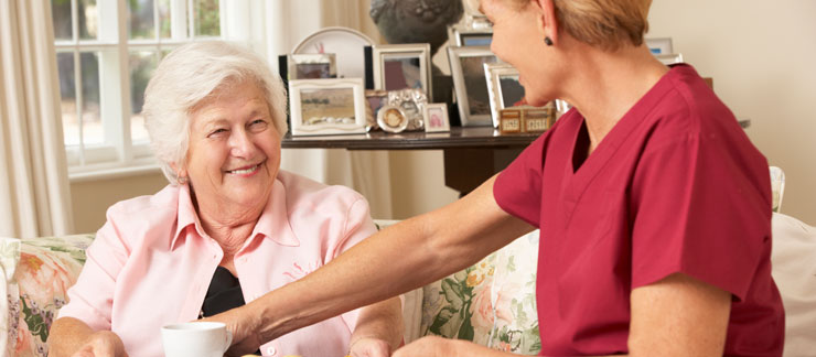 Female home care aide serves coffee to smiling senior woman sitting on a couch.