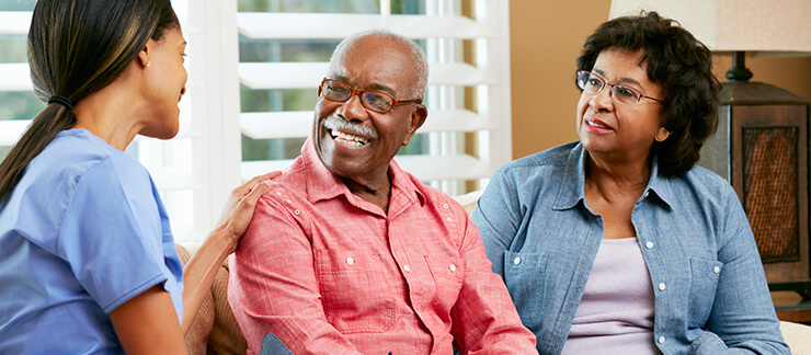 Female caregiver sits on couch while having a conversation with a smiling senior couple at home.