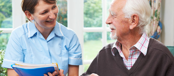 Female caregiver holding a folder smiles while talking to an elderly man at home.