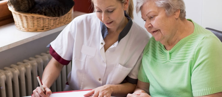 A female in-home care provider and an elderly woman in a bright green shirt sit together at a table examining a document. A cat brown cat lays on the windowsill behind them.