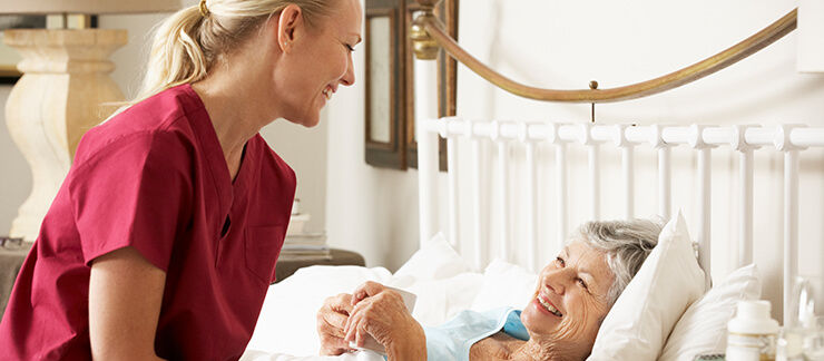 Female caregiver giving a cup of tea to smiling elderly woman lying in bed at home.