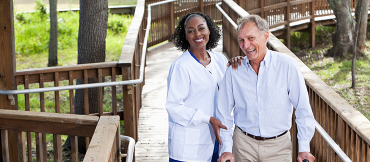 Female care aide helps senior man with walker during a stroll over a wooden bridge in park.