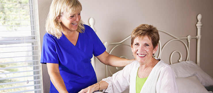 Senior woman sits on bed smiling as a female home care worker comes for a visit.