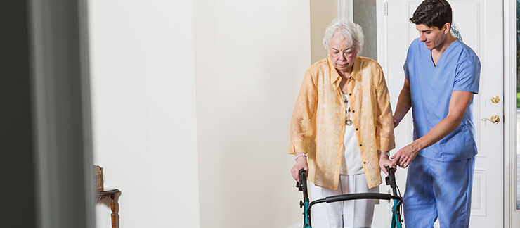 An in-home care provider dressed in blue scrubs stands next to an elderly woman in her home assisting her with her walker