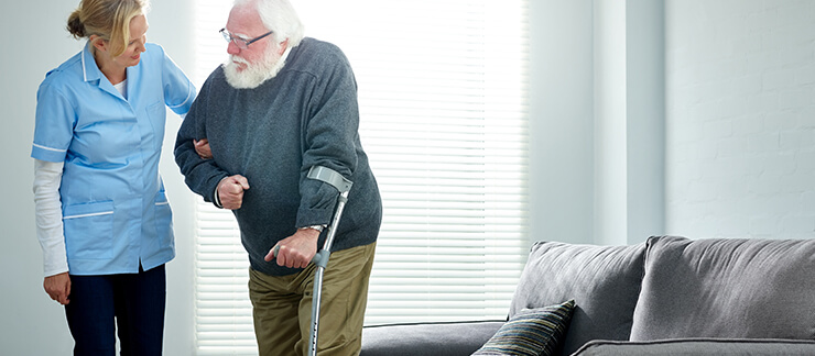 Elderly man with a cane gets help standing from couch by a helpful female caregiver.