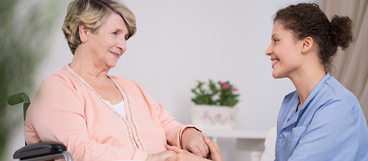 Senior woman in wheelchair holds hands with a smiling female home care aide.