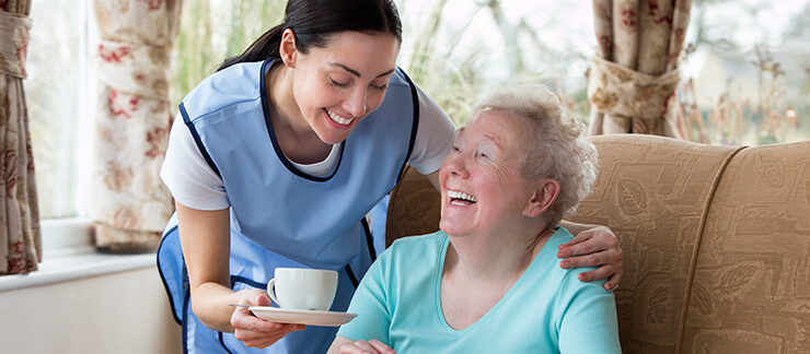 Female home care aide brings a hot cup of coffee to a smiling senior woman sitting on a couch.