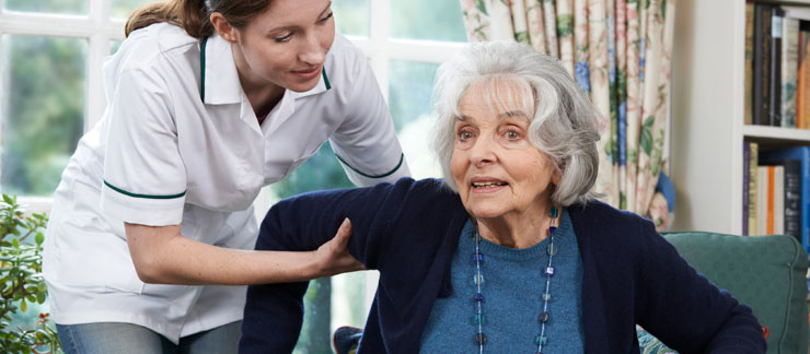 Female home care aide helps elderly woman to get up from sitting on couch.