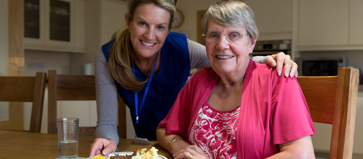 Home care provider smiles while serving breakfast to senior woman in kitchen.