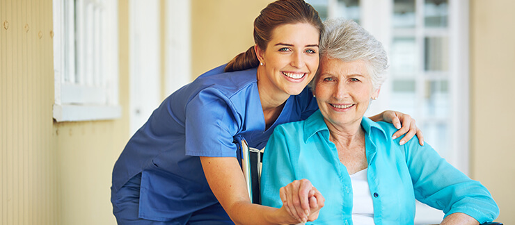 Female care provider holds hand of elderly woman sitting in front of home.