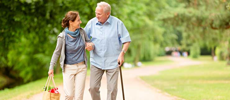 Female care aide with bag of groceries holds arm of elderly man with cane during a walk home.