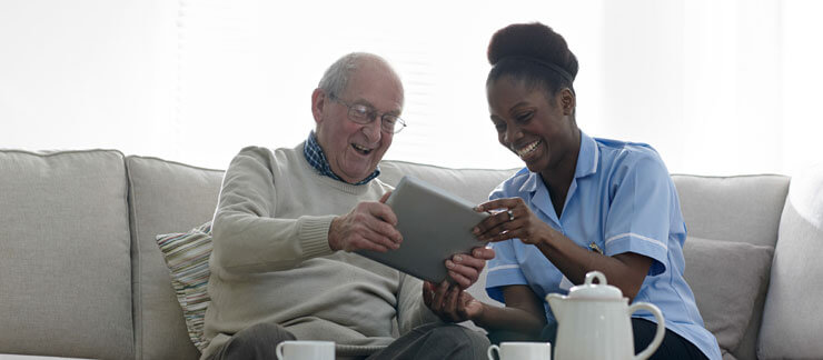 An in-home care provider and an elderly man sit side by side on a couch smiling at a tablet.