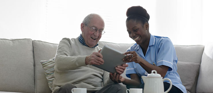 An in-home care provider and an elderly man sit side by side on a couch smiling at a tablet. There is coffee and snacks on the table in front of them.