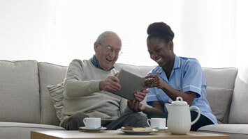 An in-home care provider and an elderly man sit side by side on a couch smiling at a tablet. There is coffee and snacks on the table in front of them.