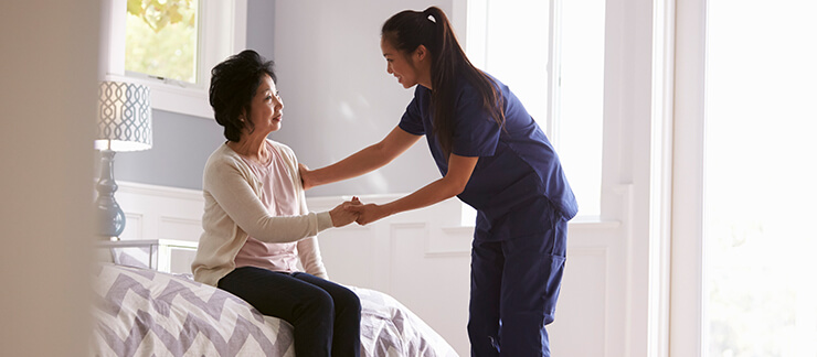 Depressed senior woman is helped out of bed by a female care aide.