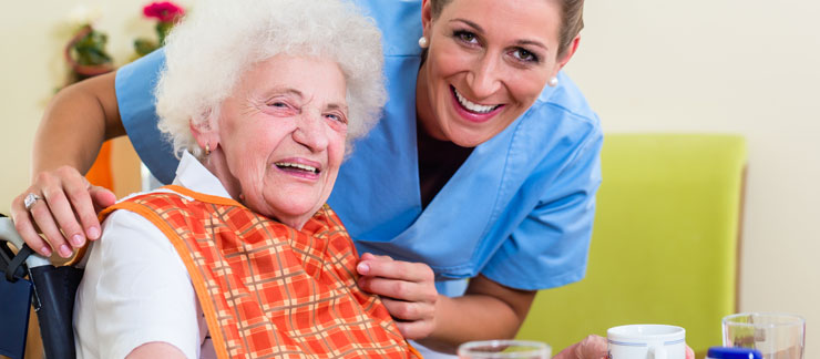 Female care worker smiles next to elderly woman sitting in a wheelchair in the kitchen.