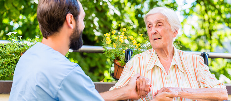 Male caregiver holds the hands of an elderly woman sitting in wheelchair outside of home.