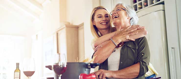 Adult daughter hugs her mother in the kitchen as they put pasta into a boiling pot of water.