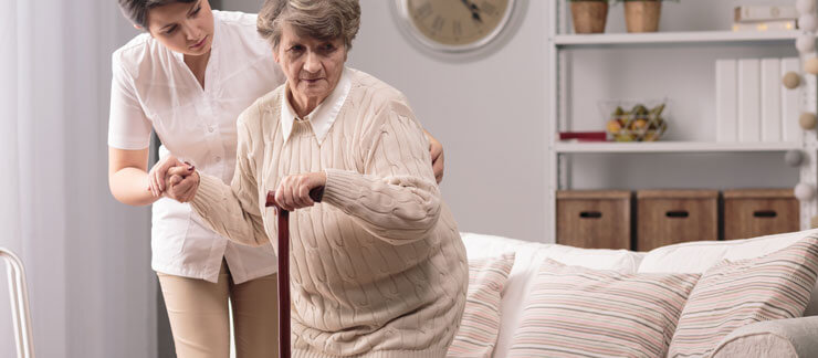 Elderly woman with cane gets up from couch with the assistance of a female home care worker.