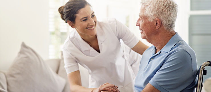 Senior man in wheelchair smiles as female care worker gently holds his hand.